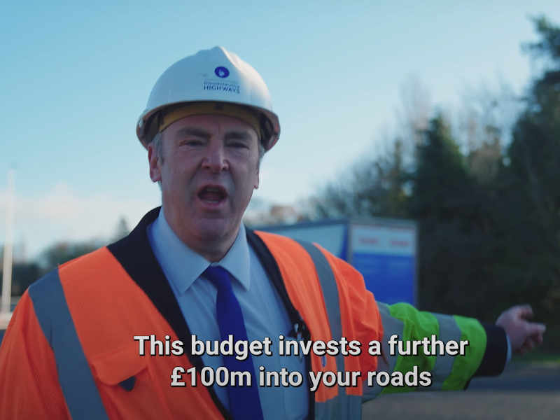 A councillor in a reflective jacket points at a lorry whilst announcing 100 million for roads in the GCC Gloucestershire budget promotional video