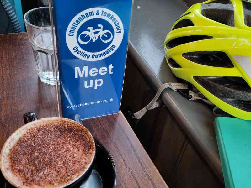 A close up of the Cheltenham & Tewkesbury Cycling Cafe meetup sign alongside a coffee and a cycle helmet.